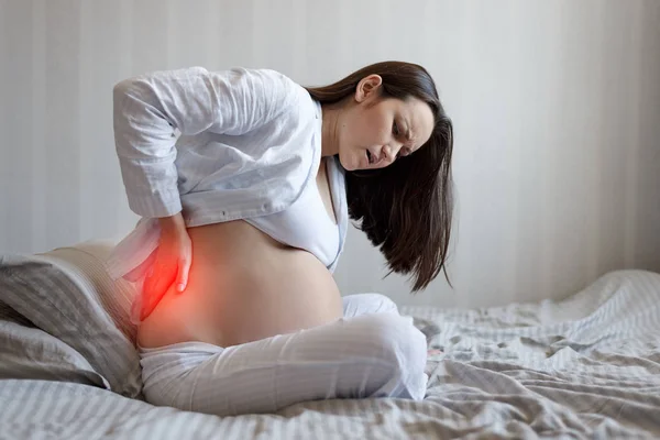 Ritratto di una donna incinta sul lato del letto che tiene la vita. mal di schiena, difficoltà di gravidanza — Foto Stock