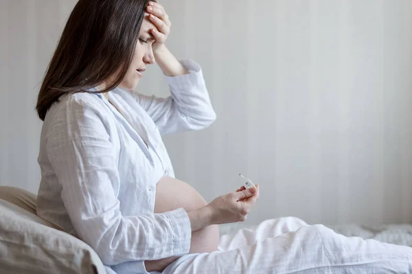 Donna incinta seduta a letto con un termometro e tenendo la testa, vista laterale — Foto Stock