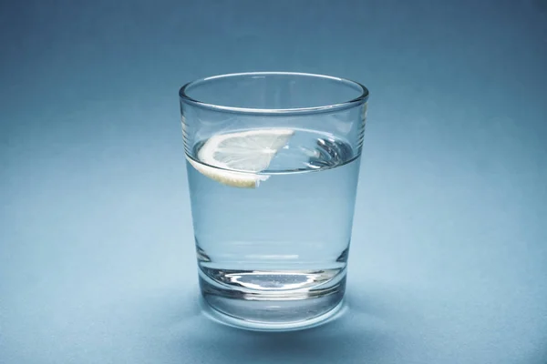 Un vaso de agua con un trozo de limón sobre un fondo azul — Foto de Stock