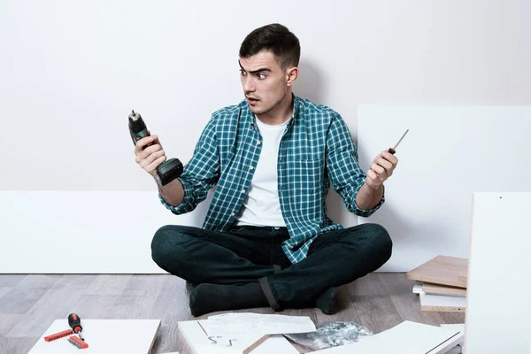 Joven desconcertado sentado en el suelo de la habitación con destornillador eléctrico y destornillador de mano, muebles Asamblea — Foto de Stock