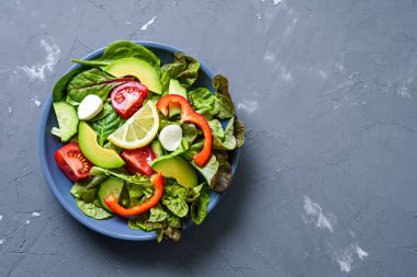 Sulu bir gri tabak üst görünümü, beton arka plan üzerinde parlak sebzeler, avokado, limon, domates, biber, salatalık ve mozzarella salatası