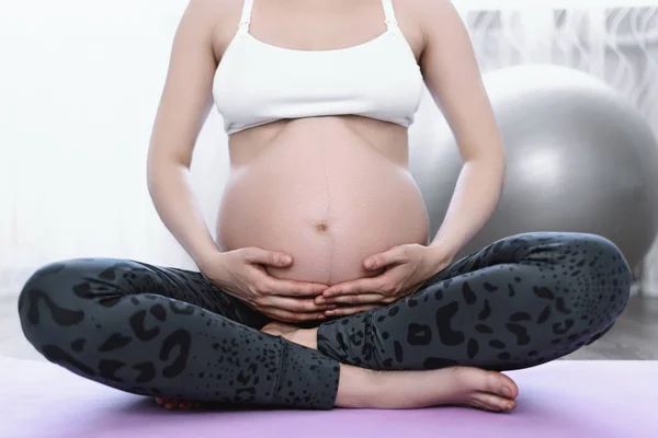 Gravid kvinna med händerna på magen sitter på sportmatta — Stockfoto