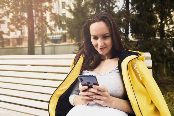Женщина в желтой куртке со смартфоном в руках сидит на скамейке — стоковое фото