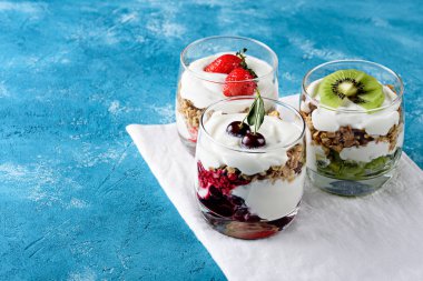 Turkuaz arka planda çeşitli berry tatlılar, granola farklı tatlılar ile üç bardak, krem şanti, çilek, kivi, kiraz