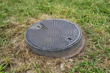 Çimenlerin ve kuru toprağın arasına yerleştirilmiş geometrik desenli yuvarlak metal kanalizasyon kapağı. Kentsel altyapı ve su ve kanalizasyon sistemleriyle ilgili kamu hizmetlerinin ayrıntıları.