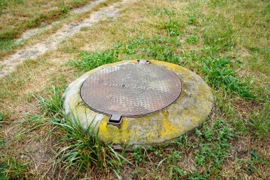 Çimenler ve kuru toprakla çevrili beton bir üsse yerleştirilmiş yuvarlak metal kaplama. Endüstriyel element ve kentsel altyapı ayrıntıları arkaplan, doku veya tasarım kavramı olarak uygun.