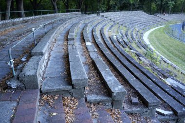 Ağaçlarla çevrili çimen ve yabani otlarla kaplı eski beton banklar çürüme ve terk edilme atmosferi yaratıyor..
