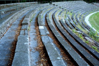 Ağaçlarla çevrili çimen ve yabani otlarla kaplı eski beton banklar çürüme ve terk edilme atmosferi yaratıyor..