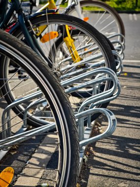 Several bicycles are parked at a metal rack under bright sunlight in a city setting. The tires are clearly visible, showcasing urban cycling culture.