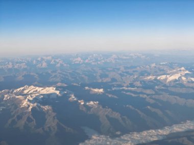 Mountains stretch across the landscape, showcasing snow-capped peaks and deep valleys under a blue sky. The scene captures the beauty from an airplane perspective on a clear day.