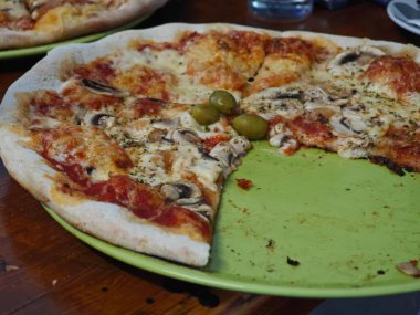 Yeni pişmiş bir pizza, üzerinde erimiş peynir, mantar, zeytin ve otlarla dolu, sıcak, davetkar bir restoran atmosferiyle çevrili yeşil bir tabakta oturur..