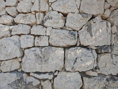 The sturdy stone wall displays various sizes of rocks, stacked unevenly, with shadows highlighting the textures. Sunlight exposes the natural weathering and earthy colors of the stones.