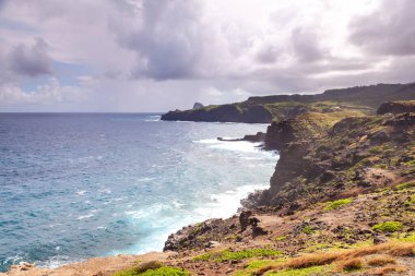 Cliffs rise dramatically along the northern coastline of Maui, where turquoise waves crash against rocky shores under a dramatic sky. 