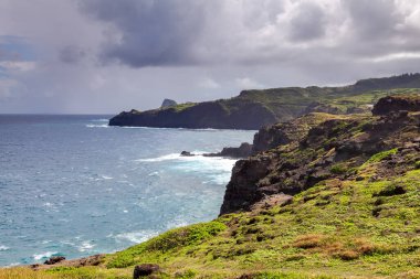 Dramatic cliffs rise above the turquoise waters of Waikeakua Gulch in the northern coast of Maui, with lush greenery contrasting against the rocky landscape under a moody sky.