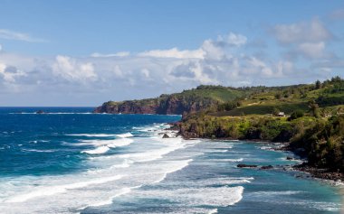 Explore the stunning north coast of Poelua Bay in Maui where waves crash against rocky shores, framed by vibrant green hills and a clear blue sky, perfect for nature lovers and adventure seekers alike.