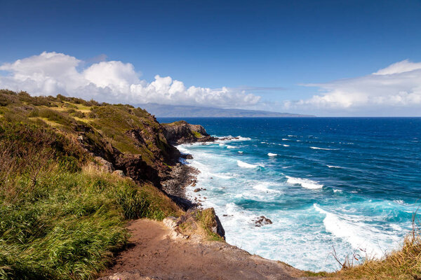 Vivid blue waves crash against the rugged northern coast around Papanalahoa Point of Maui, with green cliffs and bright skies offering stunning panoramic views.