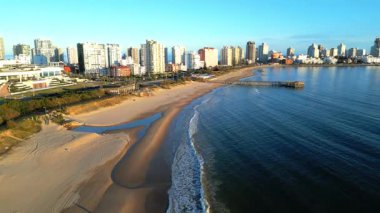 Punta del Este 'deki Playa Mansa üzerindeki İHA görüntüleri ikonik iskeleye doğru uçuyor. Rio de la Plata 'nın yumuşak dalgaları yavaşça kumlu kıyılara vururken şehir gökyüzünde yükseliyor.