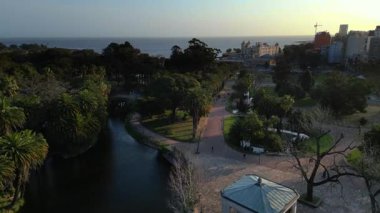 Montevideo 'daki Parque Rodo' daki drone görüntüleri, palmiye ağaçları ve yürüyüş yolları tarafından çerçevelenmiş lagünün yanındaki beyaz çardağı gösteriyor. Uçuş Mercosur binasına doğru devam ediyor.