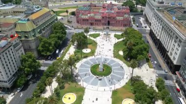 Arka planda tarihi Casa Rosada ile Buenos Aires 'teki Plaza de Mayo' nun insansız hava aracı görüntüsü palmiye ağaçları fıskiyeleri ve merkez anıtı ile geniş bir meydan dengeli bir şehir kompozisyonu oluşturur.