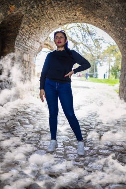 Kendine güvenen bir kadın antik bir taş kemer yolunun altında kaldırım taşı yolunda duruyor. Sisli atmosferle çevrelenmiş sahne gizemi, gücü ve zamansız mimariyi çağrıştırıyor..