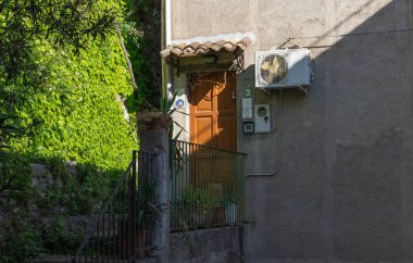 Duvara monte edilmiş bir havalandırma ünitesine bitişik küçük bir balkonda seramik tente ve saksı bitkileriyle taşralı ahşap kapı. Eski mimari ve modern hizmetler arasında büyüleyici bir tezat.