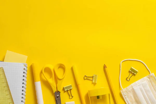 Okul malzemeleri sarı arka planda. Okul konseptine geri dönelim. Yüz için kalem, makas ve tıbbi maske. Uzayı kopyala Metin çerçevesi