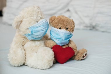 Yatağında koruyucu tıbbi maskeleri olan kırmızı kalpli iki oyuncak ayı..