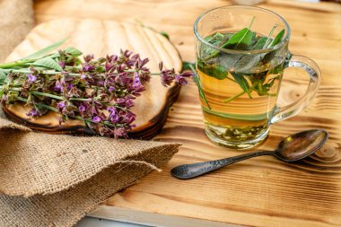 Medicinal sage tea in a glass cup with a spoon and fresh purple flowers.