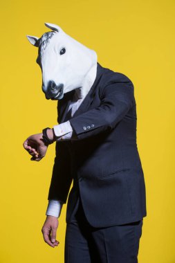A man in a suit and a horse mask looking at his watch. Yellow background. Conceptual business background clipart