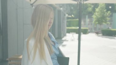 Dışarıdaki kafeye gelen ve sabah güneşinde rahatlayan bir kadın. Tarz sahibi genç bayan ceketini çıkarıp sıcak şehir terasında güne başlamak için hazırlanıyor. Yüksek kalite 4k görüntü