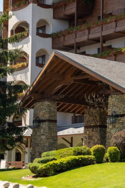 Çiçeklerle süslenmiş balkonların ve yemyeşil peyzajların bulunduğu lüks bir dağ otelinin büyük taş ve ahşap girişi. Manzaralı bir ortamda hoş geldin cephesi.