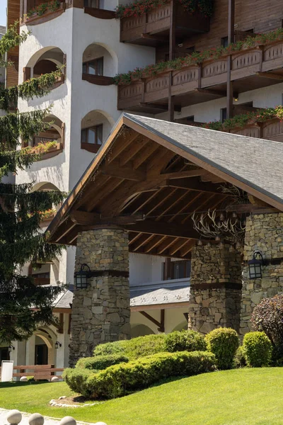 Çiçeklerle süslenmiş balkonların ve yemyeşil peyzajların bulunduğu lüks bir dağ otelinin büyük taş ve ahşap girişi. Manzaralı bir ortamda hoş geldin cephesi.