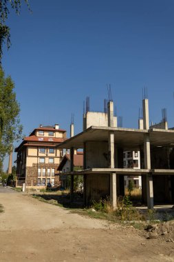 Çıplak, terk edilmiş bir beton iskelet, arka plandaki modern, tamamlanmış yerleşim yerleriyle tamamen zıt olan mavi bir gökyüzüne uzanır. Kentsel gelişim ve bozulmayı resmeder.