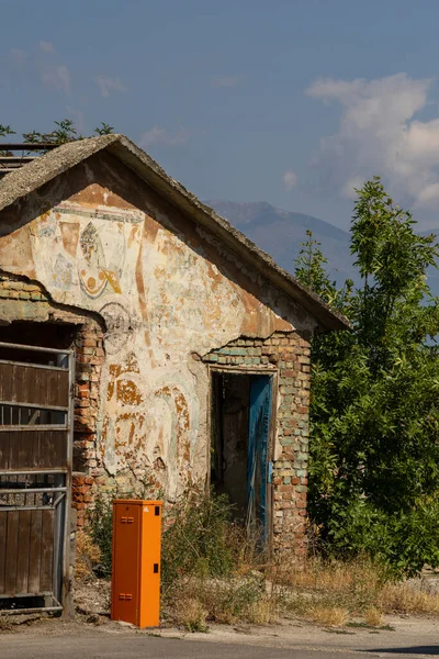 Eski, çürüyen kırsal bina, dökülen boyalar, açıkta bırakılan tuğlalar ve solmuş sanat. Ön planda parlak turuncu servis kutusu. Uzak dağlar, ağaçlar, mavi gökyüzü.