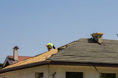 Profesyonel bir çatı ustası, açık mavi gökyüzünün altındaki bir konut binasına özenle yeni bir çatı kuruyor. İnşaat projesinde çatı malzemelerinin döşenme süreci gözler önüne seriliyor.