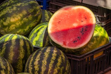 Hareketli bir pazarda canlı bir karpuz gösterisi. Biri kusursuz bir şekilde dilimlenmiş, sulu kırmızı etini ve siyah tohumlarını gün ışığıyla güzelce aydınlatıyor..