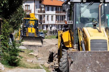 İki sarı kepçe yükleyici, güneşli bir arka planda konut binaları olan toprak bir yol kazıyor. Bu da kentsel gelişim veya altyapı çalışmalarını akla getiriyor..