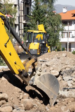 Bu görüntü, kayalık toprağı eşeleyen bir inşaat malzemesi olan sarı bir kepçe yükleyicisini yakalıyor. Olay yeri, açık bir gökyüzü altında devam eden inşaat veya kazı çalışmalarını aktarıyor.