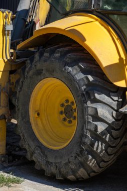 Ağır yük kepçesi yükleyicisinin engebeli tekerleği ve parlak sarı çamurluğa yakın çekim. Kirli lastik ve yıpranmış boya inşaat alanında aktif kullanıldığını gösteriyor..