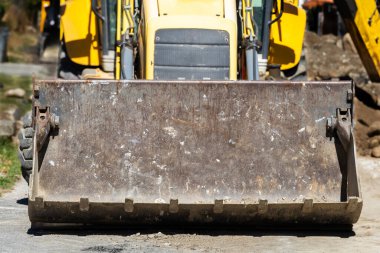 Yakın plan, yıpranmış ve kirli bir metal kovanın önü ağır bir inşaat yükleyicisine ait. Sağlam makineler dışarıda bir iş sahasında, toprak taşımaya hazır..