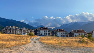 Altın tarlalar arasında modern binaların yer aldığı bir dağ beldesi manzarası. Yükselen zirveler, bazıları kar kaplı, açık mavi bir gökyüzünün altında beyaz bulutlarla yükseliyor..