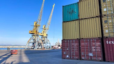 Durres Limanı, aydınlık gün. Açık mavi gökyüzünün altında iki sarı turna kuşu. Renkli istiflenmiş nakliye konteynerleri hareketli endüstriyel faaliyetler gösteriyor.