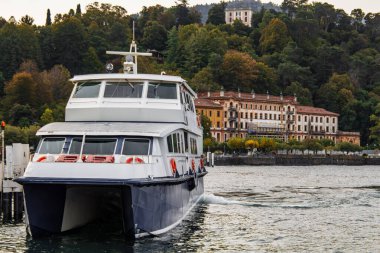 Modern katamaran feribotu Como Gölü 'nden ayrılıyor, tarihi büyük binaları ve sakin İtalya sahillerindeki gür sonbahar ağaçlarını geçiyor. Resimli manzara, klasik cazibe, sakin sular..