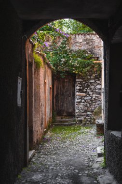 Antik taş kemer yolu, yemyeşil, canlı çiçekler, kırsal kapılar ve yıpranmış duvarlarla dolu bir kaldırım taşı ara sokağını çerçeveler. İtalya, Como Gölü yakınlarındaki Picturesque köyü..