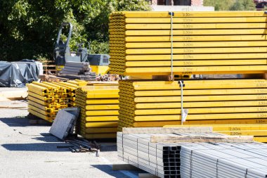 Açık hava şantiyesine yığılmış büyük sarı tahta kirişler kullanıma hazır. Arka planda çeşitli yapı malzemeleri ve ekipmanlar da görülebilir.