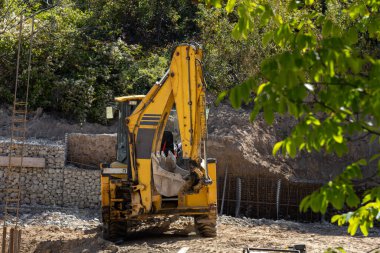 İnşaat alanındaki sarı bir kepçe yeni yerleşim yerleri için zemin hazırlıyor. Gabion duvarları ve temeller için döşenmiş inşaat demirleri erken inşa aşamasını işaret ediyor.