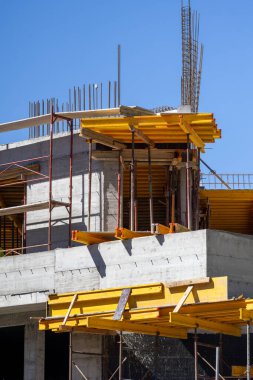 Modern bina mavi gökyüzüne karşı yapım aşamasında. Beton duvarlar, açıkta duran demirler, ve levhalar için sarı şekiller. Kentsel gelişim sürecini yakalar.
