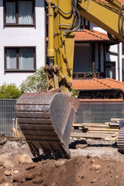 Büyük sarı kazıcı kovası yeni bir bina temeli için toprağı kazıyor. Parlak arka plandaki terasotta çatılı evler kentsel gelişimi gösteriyor.