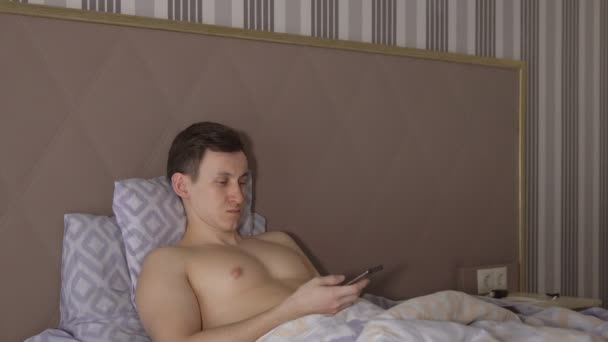 Un hombre guapo con un teléfono yace en una cama — Vídeos de Stock