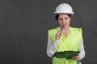 Genç kadın mühendis koruyucu kask ve yelek elinde tablet kalemiyle kemiren.